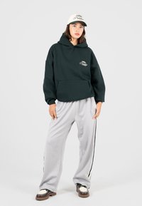 Giovane donna in piedi con felpa nera, pantaloni ampi grigi chiari con strisce nere, scarpe bianche e nere e cappellino da baseball bianco.