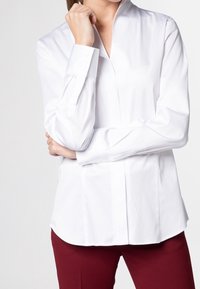 Vit skjorta med knappstängning, långa ärmar och rak skärning; bärs med vinröda byxor. Slät och krispig tygtextur.