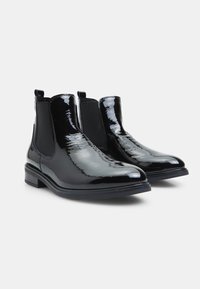 Botines de charol negro con paneles laterales elásticos, puntera redonda, tacón bajo y tira de sujeción para poner y quitar con facilidad. Textura suave.
