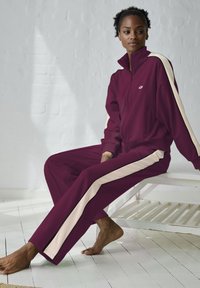 Bordo trenirka s kremastim bočnim prugama, koja uključuje jaknu na zip i odgovarajuće hlače. Glatka tkanina, opušteni kroj i minimalistički dizajn.