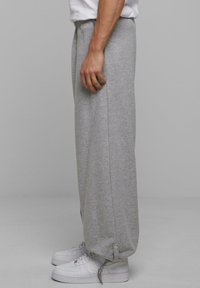 Grå sweatpants med løs pasform, elastisk talje og sidelommer. Har en knaplukning ved anklen. Bæres med hvide sneakers.