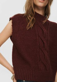 Moteris vilki be rankovių, bordo spalvos megztinį su pynių raštu ir banguota apatine dalimi, kartu su mėlynomis džinsinėmis kelnėmis ir auksinėmis apyrankėmis.