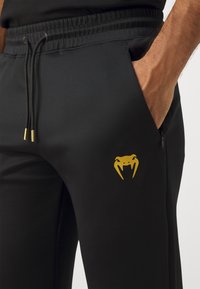 Črne joggers hlače iz gladkega materiala z elastiko v pasu, ki imajo zlate zankaste logotip na stegnih in zadrgo na stranskih žepih.