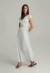Robe longue blanche à pois noirs, avec des manches volantées, un design croisé et un nœud à la taille. Associée à des sandales noires à lanières.