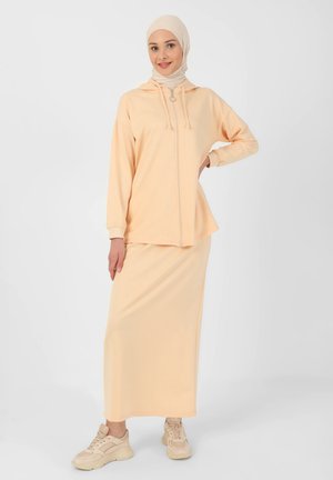 Peach-colored loungewear set featuring a zip-up hoodie and a matching long skirt. Soft material with a relaxed fit, complemented by beige sneakers.