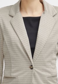 Blazer beige clair à motif à carreaux, avec des revers crantés, une fermeture à un bouton et deux poches avant. Porté sur un haut noir.