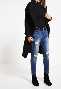 Cappotto lungo nero su un top nero, abbinato a jeans blu strappati e stivali neri alla caviglia. L'outfit presenta un design elegante e una vestibilità casual.