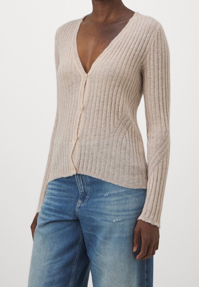 Cardigan beige clair côtelé avec un décolleté en V profond, manches longues et bords légèrement effilochés. Comprend des boutons sur le devant et une coupe ajustée.