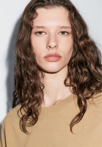 Nahaufnahme einer Person mit langen, welligen braunen Haaren, die ein hellbraunes Rundhalsoberteil trägt. Der Hintergrund ist ein sanftes Grau.