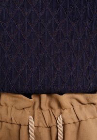 Suéter de punto en azul oscuro con un patrón de diamantes sobre una tela color beige que presenta acentos agrupados y un cordón ajustable.