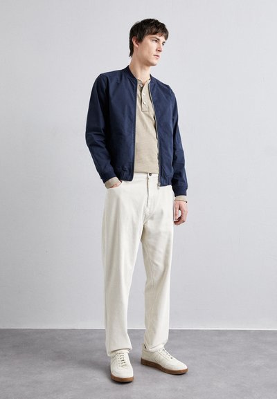 Chaqueta bomber azul marino, camiseta henley beige, pantalones de pana blancos y zapatillas blancas con detalles beige. Diseño minimalista, corte casual.
