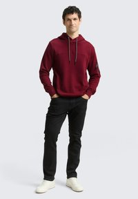 Felpa bordeaux con cappuccio e cordini regolabili, design testurizzato e tasca con zip sulla manica sinistra, abbinata a jeans neri e sneakers bianche.