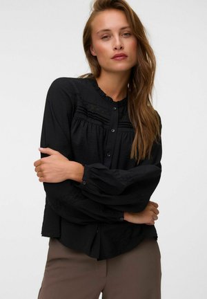 Femme aux longs cheveux bruns portant un chemisier noir boutonné avec un col à volants et un pantalon marron, posant les bras croisés devant un fond uni.
