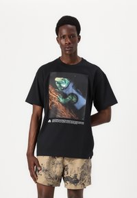 SchwarzKurzarm-T-Shirt mit einem bunten Iguana-Motiv und Text darunter. Weicher Stoff; lässiger Schnitt. Kombiniert mit gemusterten hellbraunen Shorts.
