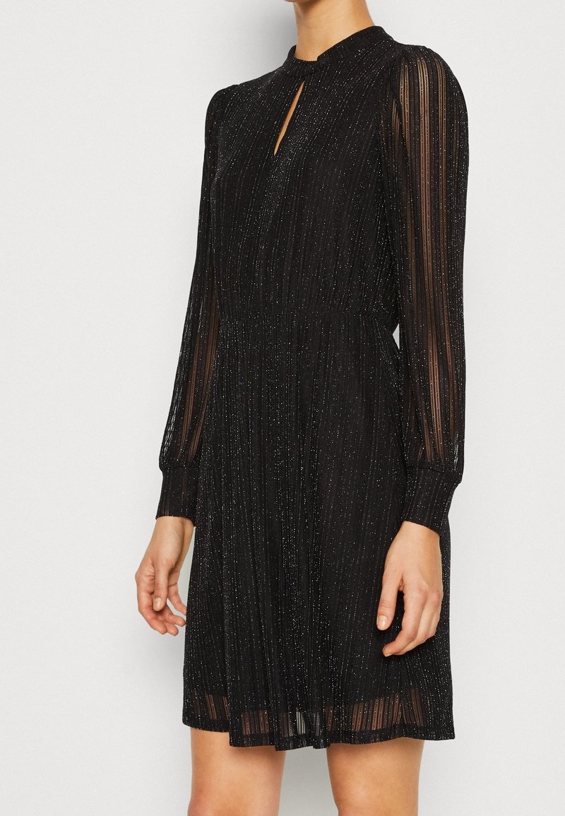 Femme portant une robe noire à manches longues avec des rayures verticales transparentes et un subtil éclat argenté, encolure en trou de serrure et jupe plissée.