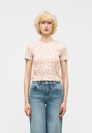 Donna con capelli biondi ricci che indossa un top corto a maniche corte rosa pallido con piccoli motivi floreali e jeans a vita alta azzurri.