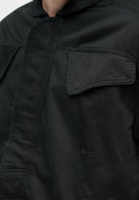 Fekete dzseki tartós anyagból, két első zsebbel, gallérral és finom márkajelzéssel a bal mellkason. Simaságú textúra.