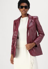 Blazer in similpelle bordeaux con una finitura liscia, revers a(gi)/notch e tasche frontali, abbinato a un maglione leggero e una gonna con frange.