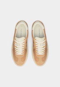 Ζευγάρι μπεζ και κρεμ αθλητικά παπούτσια με λευκά κορδόνια, φωτογραφημένα από πάνω σε ανοιχτό φόντο.