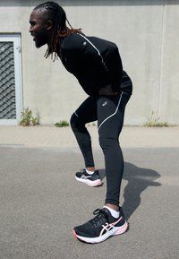 Top atlético negro con detalles en blanco, leggings negros ajustados y zapatillas de correr negras con detalles en rosa. Entorno urbano al aire libre, mostrando movimiento.