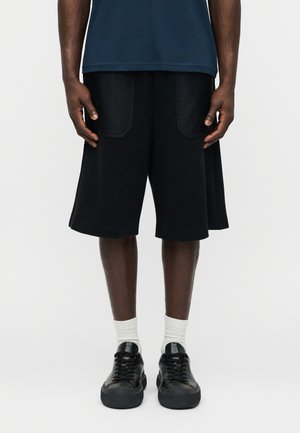 Hombre con pantalones cortos negros hasta la rodilla con bolsillos delanteros, calcetines blancos, zapatillas negras y una camisa de manga corta azul oscuro, de pie sobre un fondo blanco.