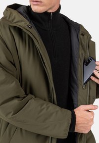 Olivově zelená bunda s kapucí, se zipem vpředu a bočními kapsami. Černý pletený rolák pod ní. Chytrý telefon v vnitřní kapse.