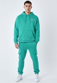 Turkis hoodie med frontlomme, snoretræk og logo, parret med matchende joggers med cargo-lommer og ribbede manchetter. Hvide sneakers.