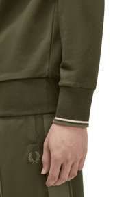 Sudadera y pantalones verde oliva, puños acanalados con un acento blanco, logo bordado en los pantalones, textura suave, diseño ajustado.