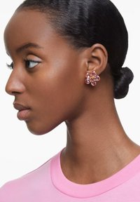 Różowe kolczyki w kształcie kwiatów z wieloma płatkami i centralnym kamieniem w odcieniach różu, noszone przez modelkę o gładkiej, ciemnej karnacji.