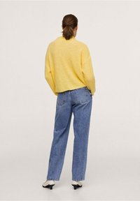 Hellgelbes, gestricktes Sweatshirt mit lockerer Passform, kombiniert mit hochtaillierten blauen Jeans und weißen, hochhackigen Mules, von hinten betrachtet.