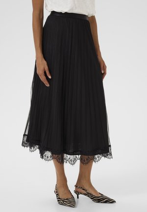 Femme portant une jupe midi plissée noire avec une finition en dentelle et des escarpins à talons kitten au motif zèbre, se tenant devant un fond blanc uni.