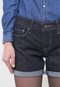 Pantalones cortos de mezclilla oscura con dobladillos enrollados, que cuentan con cinco bolsillos, cierre de botón metálico y costuras amarillas contrastantes contra una camisa de chambray azul.