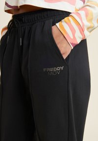 Pantalones de chándal negros con cintura ajustable, bolsillos laterales y un logo plateado "FREDDY MOV" en el frente. Tejido suave, corte relajado.