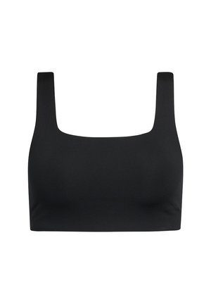 Woman wearing black crop top, black jogger pants, and silver sneakers, adjusting waistband, standing on plain light background.