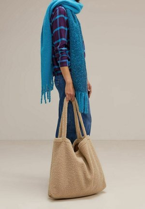 Personne portant une chemise à carreaux bleus et un jean tenant un grand sac fourre-tout beige texturé, enveloppé dans une épaisse écharpe bleue sur un fond uni.