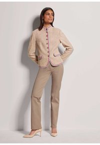 Beige strukturierte Jacke mit hohem Kragen und kontrastierendem pinken Besatz, mit vorderen Knöpfen und zwei Taschen. Kombiniert mit beigen Hosen und Schuhen mit hohen Absätzen.