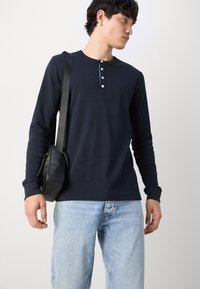 Marineblå langermet henley-skjorte med fem-knapps placket, sammen med lyseblå jeans og en liten svart crossbody-veske.