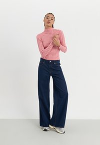 Rdeče-bele prugaste turtleneck majice, v paru z temno modrimi širokimi hlačami. Opazne detajle vključujejo visok ovratnik in subtilno šivanje.