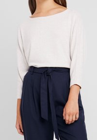 Haut beige clair à manches longues avec un large décolleté, associé à un pantalon taille haute bleu marine foncé avec un détail de ceinture à nouer. Texture douce.