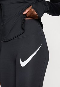 Svarta träningsleggings med en framträdande vit Nike swoosh-logotyp, slät textur och hög midjebandsdesign.