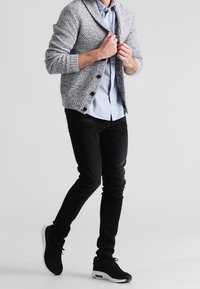 Szary dzianinowy kardigan z guzikami, jasnoniebieska koszula, czarne dopasowane jeansy i czarne trampki. Materiał o fakturze, luźny krój, codzienny styl.