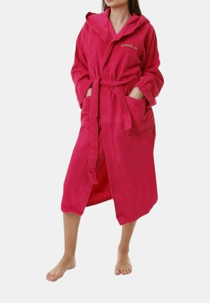 Donna con un accappatoio con cappuccio rosa brillante con tasche frontali e cintura, in piedi a piedi nudi su uno sfondo semplice.