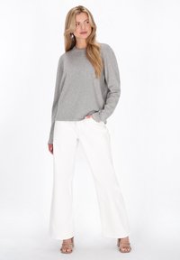 Grijze geribbelde trui met lange mouwen, gecombineerd met witte broek met wijde pijpen en nude hakken. Model staat zelfverzekerd tegen een effen achtergrond.