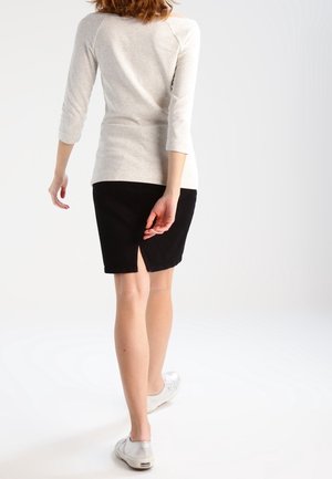 Vrouw loopt weg en draagt een lichtgrijze top met lange mouwen, een zwarte kokerrok tot op de knie met een split aan de achterkant en witte sneakers tegen een witte achtergrond.