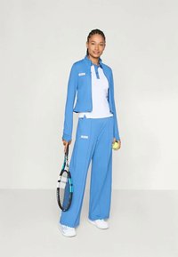 Blue athletic outfit featuring a fitted jacket with a high collar, white polo underneath, loose blue pants, and white sneakers, holding a tennis racket.