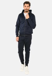 Felpa con zip color blu navy e cappuccio con cordino, abbinata a pantaloni jogger coordinati. Tessuto morbido, polsini a coste e sneaker completano il look.