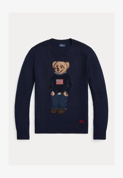 Tamsiai mėlynas megztinis su meškiuku, dėvinčiu džinsus ir juodą megztinį su Amerikos vėliava, raudonos „RL“ inicialai apačioje.