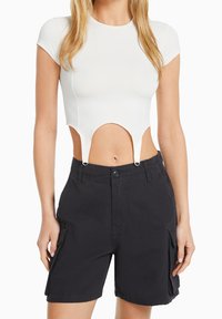 Bílý crop top s krátkými rukávy, který se vyznačuje unikátním výřezem a kovovými detaily. V kombinaci s černými cargo šortkami.