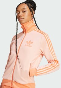 Adidas trenirka svijetlo ružičaste boje s narančastim naglascima, visokim ovratnikom, zatvaranjem na zip i tri pruge na rukavima. Mekana, glatka tkanina.