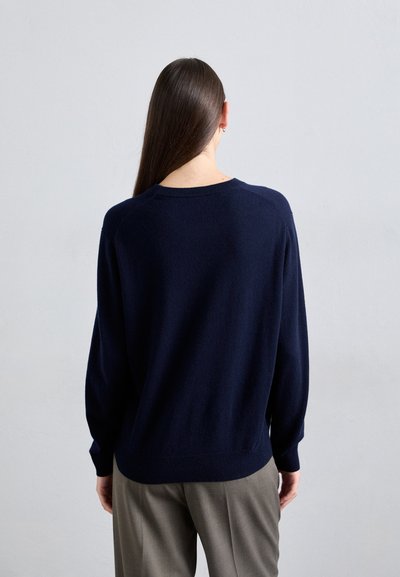 Person with long straight hair wearing a navy blue sweater and gray pants, standing facing a plain light gray wall.
